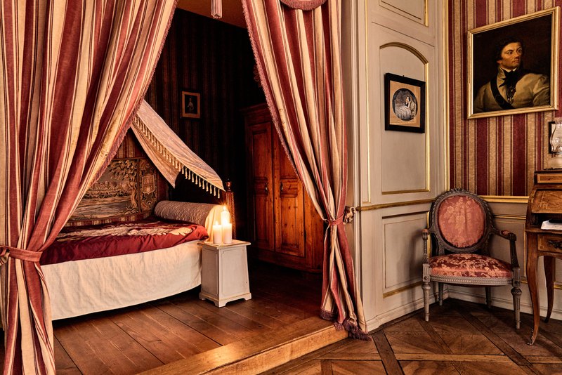 Kościuszko’s bedroom at the Zeltner house, Gurzelngasse 12 in Solothurn — a canopied bed in crimson-and-gold fabric, a burning candle on the bedside table, a portrait of Kościuszko on the wall beside the alcove. The room has been preserved since 1817 and is today the central exhibit of the Kosciuszko-Museum Solothurn.